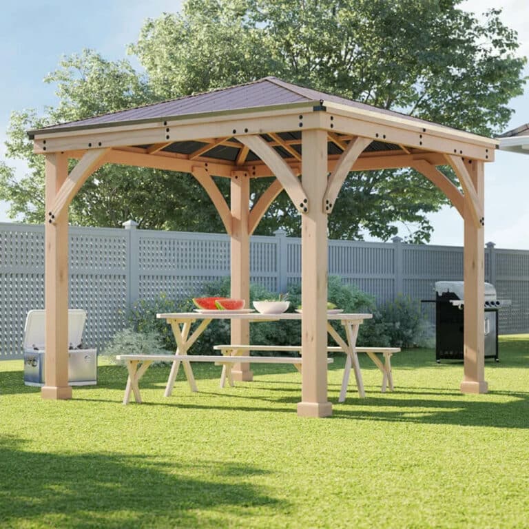 Backyard scene featuring a wooden gazebo with a pitched roof, picnic table, and benches, set on a grassy lawn with a barbecue grill and fencing in the background.