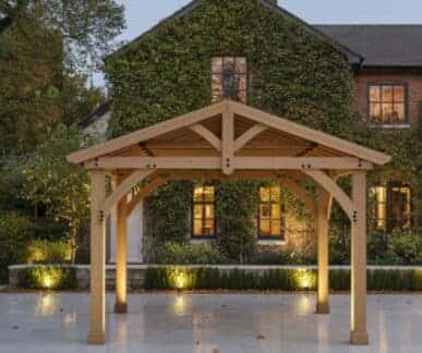 Wooden gazebo with a gabled roof and diagonal braces, set on a polished stone patio in front of an ivy-covered brick house with illuminated windows, surrounded by trimmed hedges and autumn trees under dusk lighting.