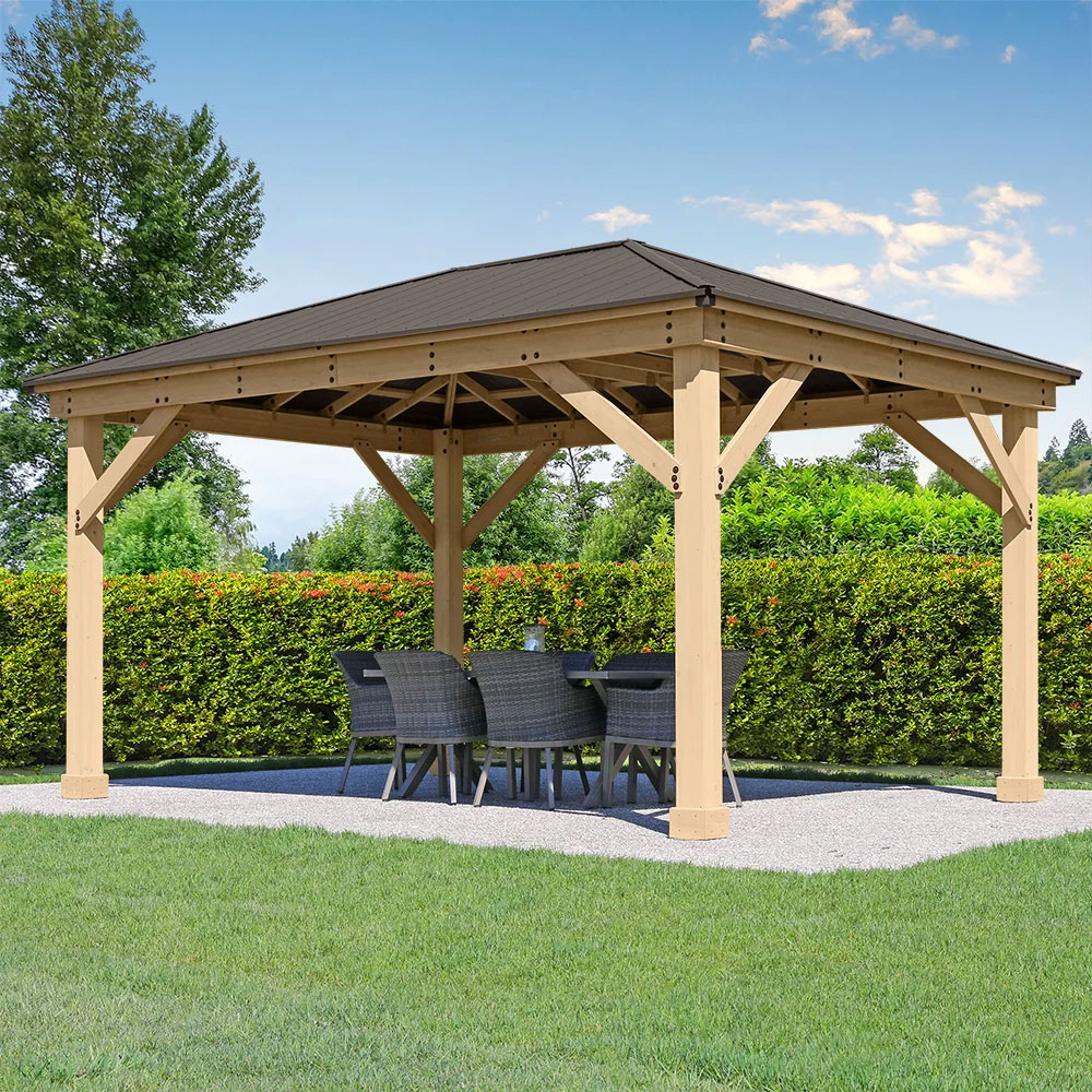 A photo of a modern outdoor wooden gazebo with a dark brown, pitched roof, set up on a white gravel patio area. The gazebo has thick, light-colored wooden posts and structural beams. Under the shelter, there is a dark rectangular outdoor dining table surrounded by six woven, gray-colored chairs. The gazebo sits on a neatly trimmed patch of grass, and behind it is a tall, dense green hedge, with trees visible in the background under a bright blue sky with some light clouds.