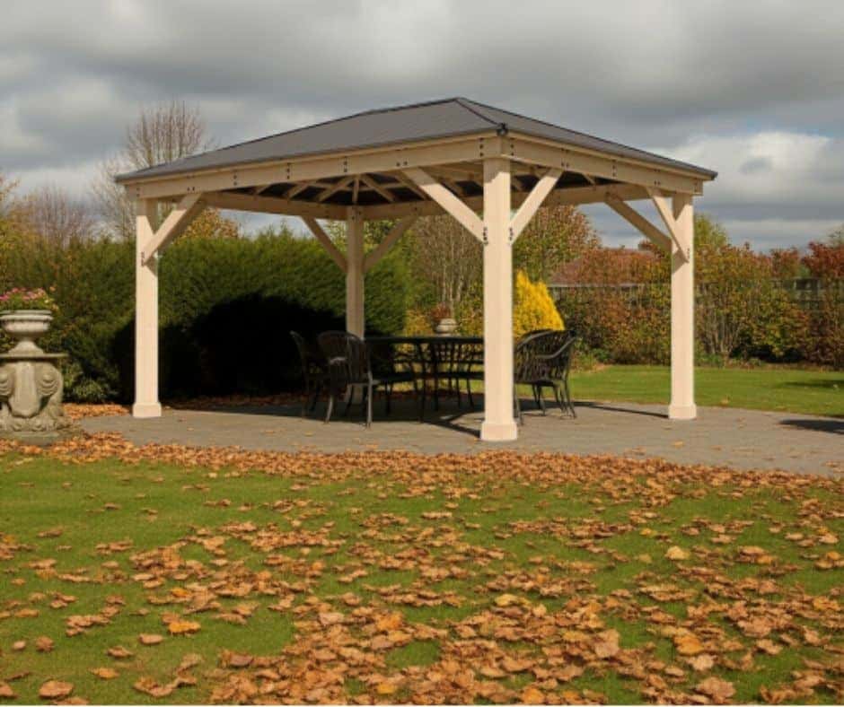 Wooden gazebo with a dark pitched roof supported by four timber posts, sheltering a dining table and chairs on a paved patio, surrounded by trimmed bushes, trees with autumn foliage, and fallen leaves on the grass under a partly cloudy sky.