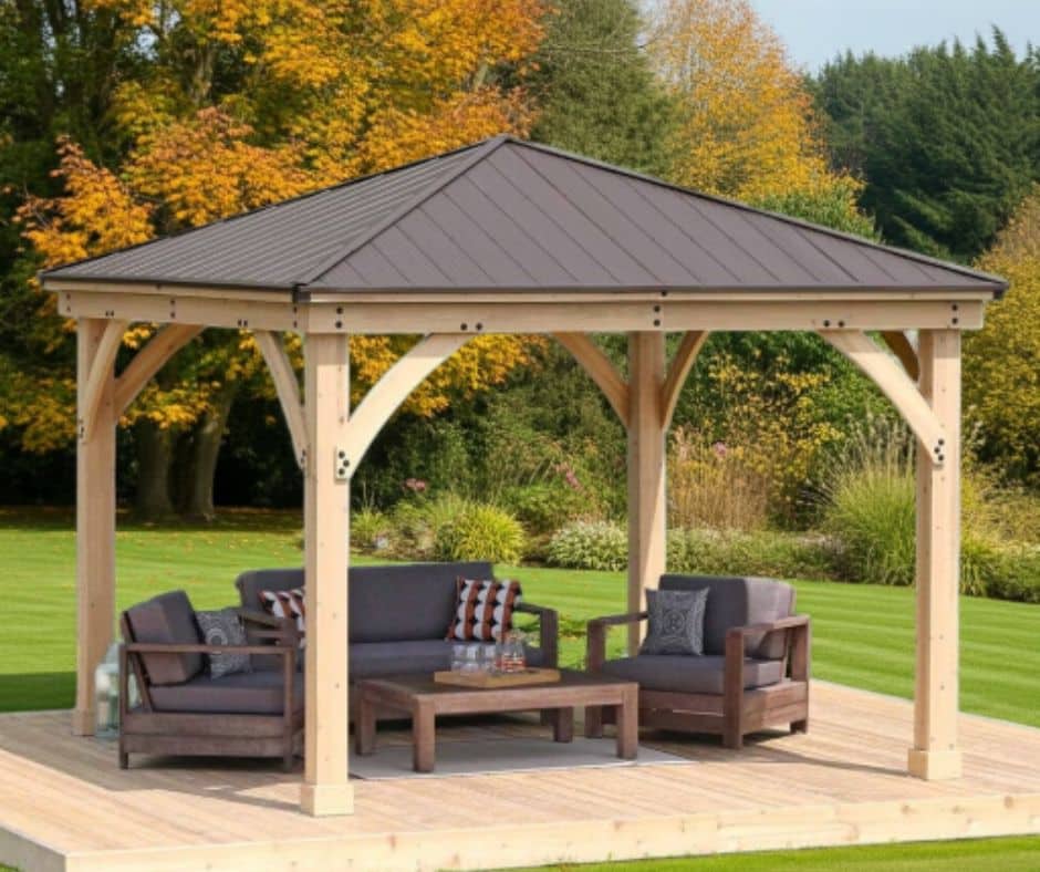 Wooden gazebo with a dark metal pitched roof, featuring two armchairs and a sofa with dark upholstery, arranged around a wooden coffee table on a raised wooden platform, set in a landscaped garden with green lawn and autumn trees in the background.