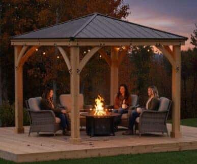 Wooden gazebo with a metal roof and string lights, featuring wicker armchairs around a lit firepit, with three people seated and autumn trees in the background.