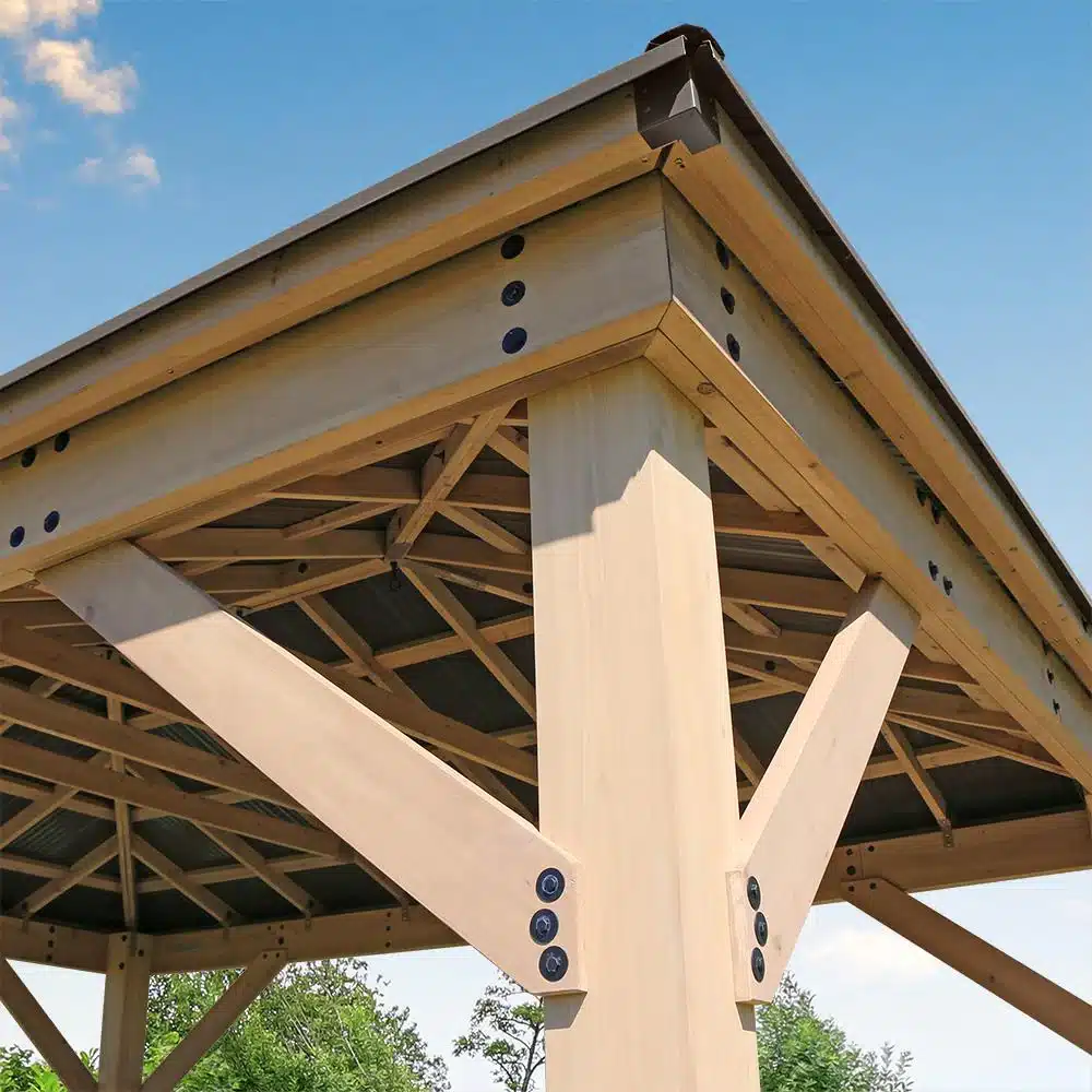 Corner detail of a wooden gazebo with pitched roof, showing beams, braces, and metal bolts against a blue sky and green foliage.