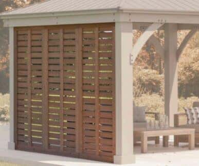 Modern outdoor gazebo with a metal roof and slatted wooden privacy wall, featuring cushioned seating and a wooden coffee table in a landscaped garden.