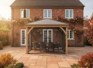 Wooden gazebo with a metal roof, featuring a round table and six wicker chairs on a stone patio, set against a brick house with climbing white roses and surrounded by shrubs and plants.