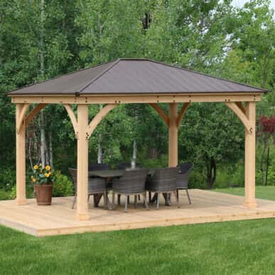 Wooden gazebo with a dark metal roof sheltering a dining table and six woven chairs on a raised timber deck, surrounded by lush greenery and trees.