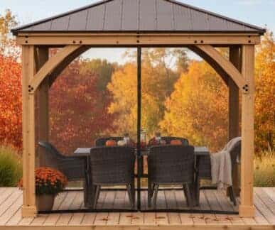 Wooden gazebo with a metal roof on a deck, featuring a rectangular dining table with pumpkins, glassware, and an autumn centre piece, surrounded by wicker chairs and autumn foliage. Mesh curtains cover the sides.