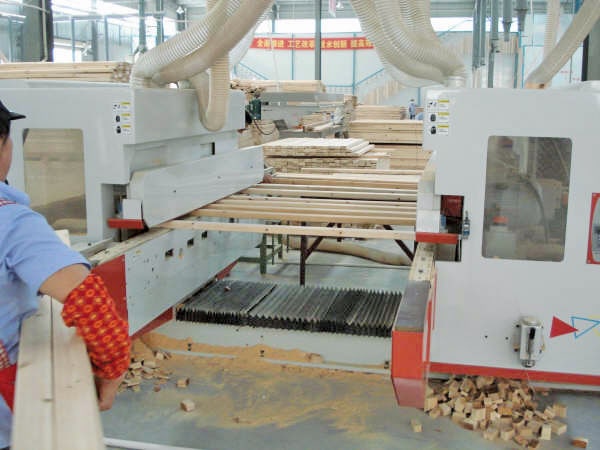 Woodworking factory with automated machine processing wooden planks, surrounded by sawdust and monitored by a worker in protective gear.