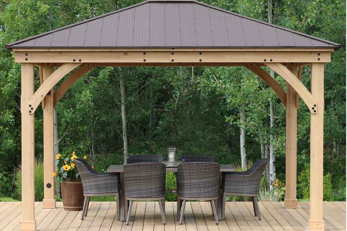 Yardistry Wooden Gazebo 14ft x 12ft (4.3 x 3.7m), Meridian Series 4.3m x 3.7m Wooden Gazebo Premium Cedar Gazebos with Aluminium Roofs Wooden gazebo with a dark metal roof sheltering a rectangular dining table and six wicker chairs on a timber deck, set against a lush forest backdrop.