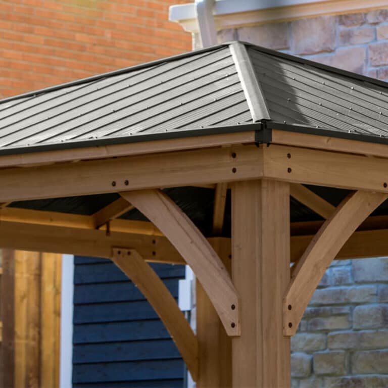 Close-up of wooden gazebo with dark metal roof and curved braces, set near stone and brick walls in a backyard setting.
