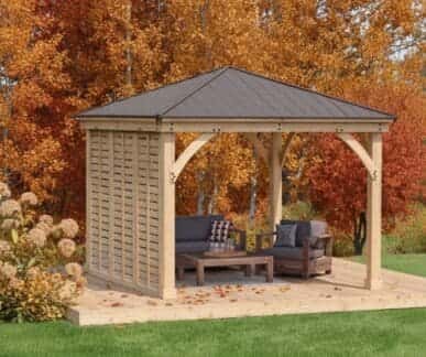 Wooden gazebo with a dark metal roof and vertical slatted privacy wall, featuring a cushioned sofa, armchair, and coffee table on a wooden deck, surrounded by autumn trees and scattered leaves.