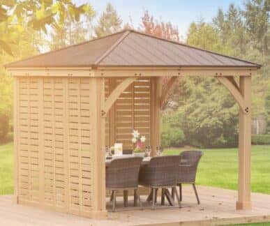 Wooden gazebo with a dark metal roof and slatted walls, featuring a rectangular dining table with a white tablecloth, floral centerpiece, and four wicker chairs, set on a wooden deck surrounded by grass and autumn trees.