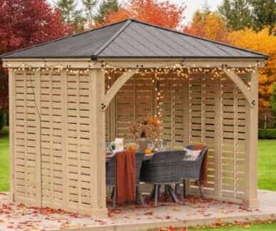 Wooden gazebo with a black metal roof and string lights, featuring a dining table with a burnt orange tablecloth, woven chairs, and a dried flower centerpiece, set on a wooden deck surrounded by autumn trees and fallen leaves.
