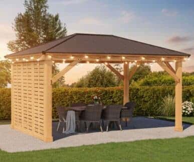 Wooden gazebo with a dark pitched roof and string lights, featuring a round dining table with six wicker chairs and a floral centerpiece, set on a gravel patio surrounded by lawn and hedges.