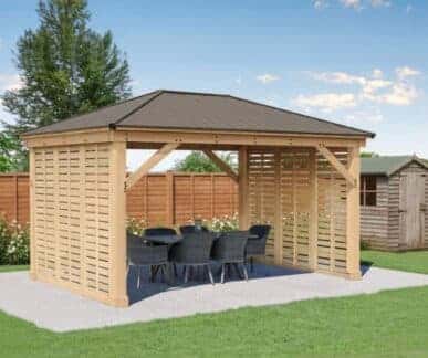 Wooden gazebo with slatted walls and a dark pitched roof, featuring a dining table with eight black woven chairs, set on a concrete pad in a landscaped garden with green lawn and autumn foliage.