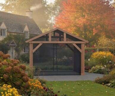 Wooden mocha gazebo with a gabled roof and mesh kit enclosure, surrounded by blooming autumn flowers and set in a landscaped garden with a stone cottage in the background.
