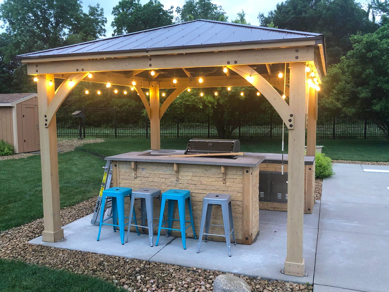 Outdoor kitchen and bar under wooden pergola with metal roof, string lights, built-in grill, stone countertop, and colourful metal bar stools on a paved patio.