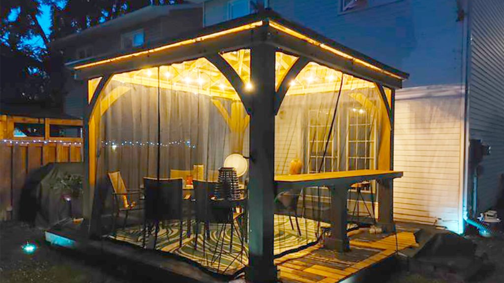 Wooden gazebo with shelf, framed with mosquito mesh curtains that are drawn to offer protection.  Low level lighting provides ambiance, showcasing the wicker and glass furniture design for relaxing and entertaining.  A hurricane lamp sits atop a wood and metal stool in the background.  String lights adorn the fence in the background and the house at the rear appears white in colour.