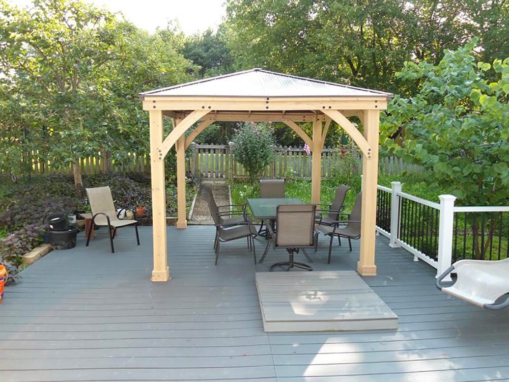 Timber gazebo with metal roof on gray-painted deck featuring glass-top table, six chairs, and garden backdrop