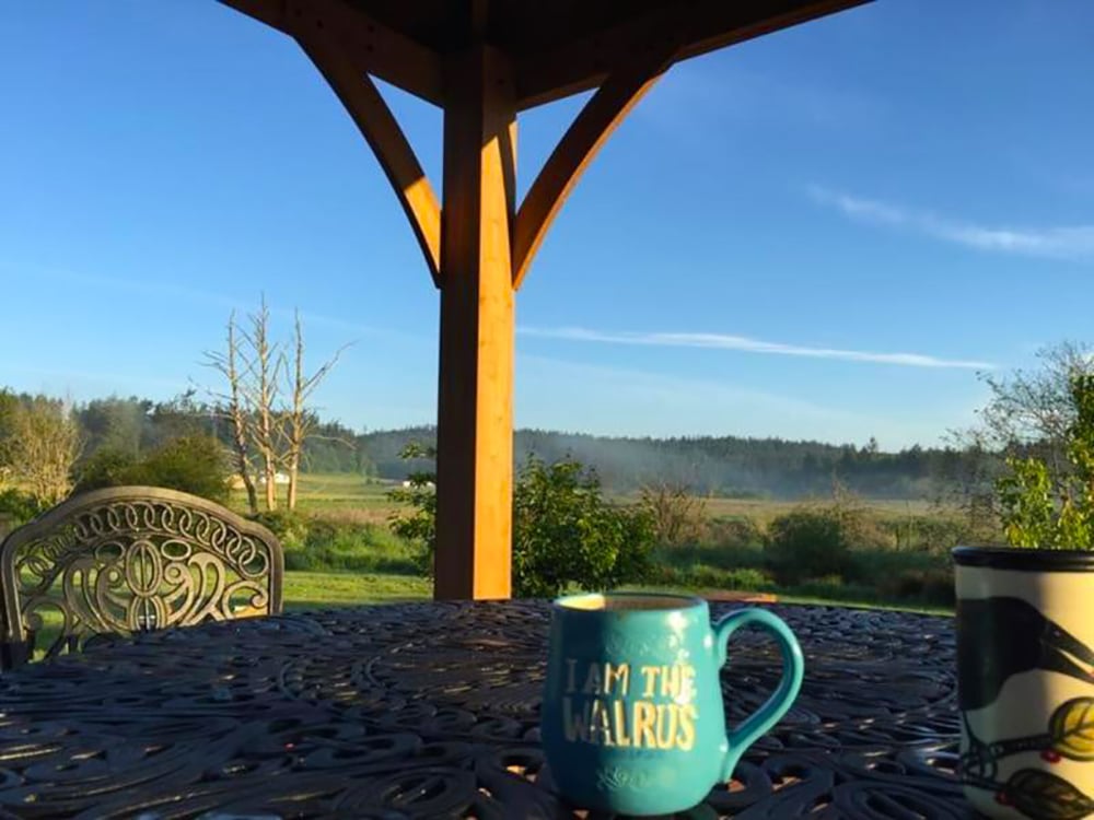 This image presents a serene outdoor morning scene, focusing on a table setting with a broad, expansive view of the countryside. In the foreground, a dark, cast-iron or intricately detailed metal outdoor table fills the lower half of the frame. A portion of a matching outdoor chair with an ornate backrest is visible on the left.

The central focus is a bright teal-blue ceramic mug resting on the table. In bold, gold lettering, the mug bears the popular phrase,