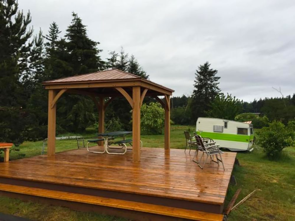 A photo of a newly installed, large wooden deck with a gazebo in a grassy outdoor area, likely a park or campground, on an overcast day. The deck appears wet and has a rich, glossy brown finish. A wooden gazebo with a copper-colored, metal hip roof stands on the deck, sheltering a traditional green metal picnic table and benches. Several lightweight metal chairs are also placed on the open deck area. In the background to the right, a vintage white and green travel trailer is visible amidst the trees and brush. Tall evergreen trees dominate the background under a gray sky.