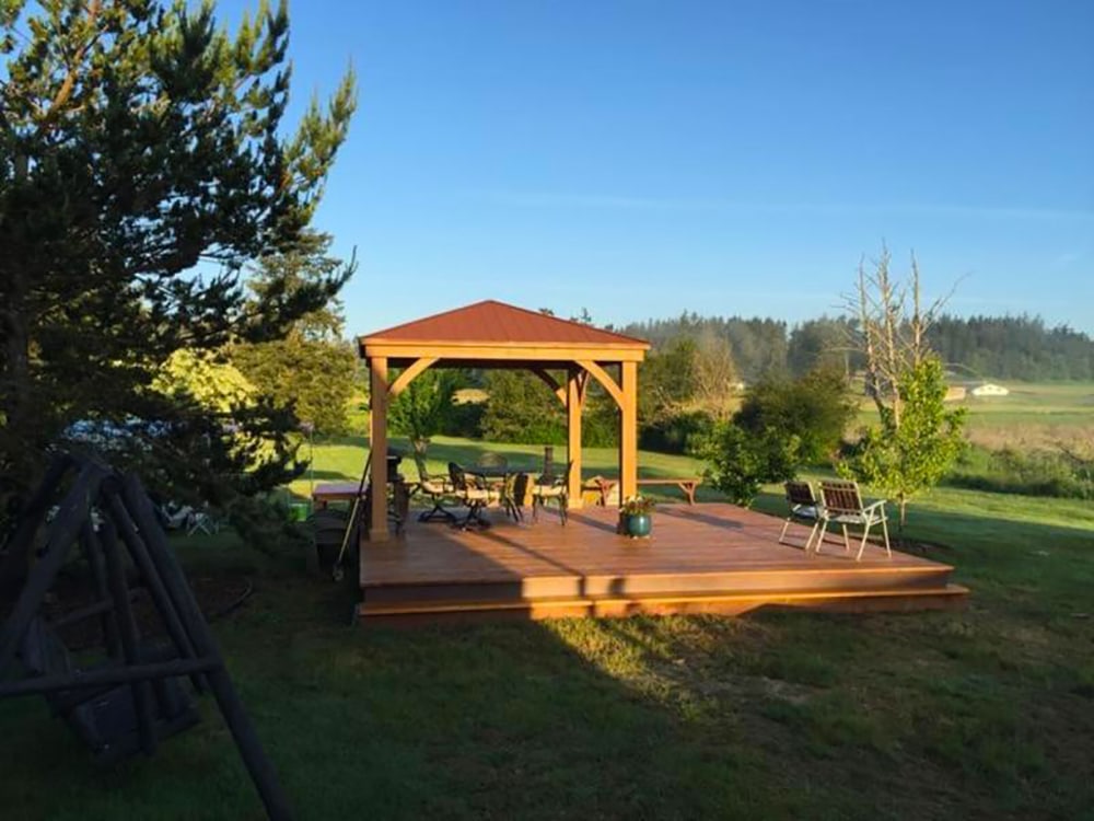 A bright, wide-angle photo of an outdoor wooden deck and gazebo setup in a grassy, rural setting. The rectangular wooden deck, elevated with a step, features a gazebo with a warm, reddish-brown roof and a wooden frame centered on the deck. Under the gazebo, there is an outdoor dining set with chairs. The deck is surrounded by lush green grass and various trees and foliage. To the left, a dark-colored swing set frame is partially visible. In the background, the landscape opens up to a large field with distant trees and a few small houses under a clear blue sky.