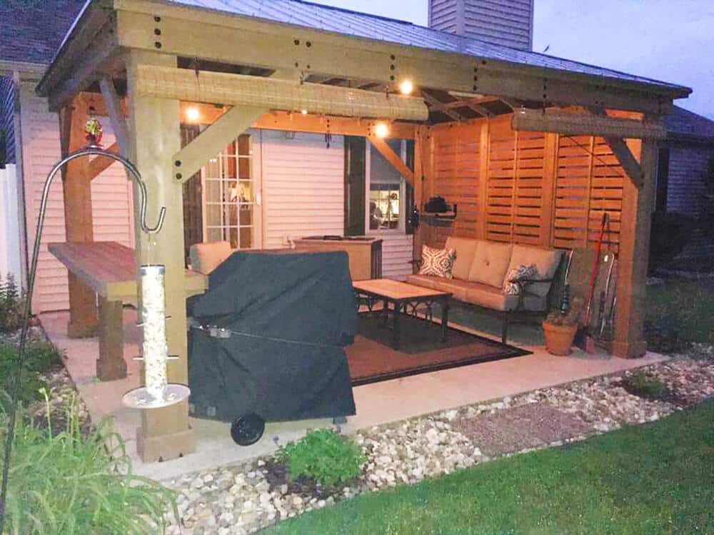 A warm, ambient evening shot features a well-lit wooden patio gazebo attached to the side of a suburban house. The photo is taken at dusk, with the exterior lights creating a cozy atmosphere.

The gazebo structure is made of light brown stained wood with a slatted metal roof and partially enclosed sides using horizontal wooden screens, visible on the right. String lights with exposed bulbs are suspended along the inner perimeter of the roof, providing soft, inviting illumination. Bamboo roll-up blinds are partially lowered beneath the roof eaves, adding a decorative touch and extra shade or privacy.

The patio underneath the gazebo is concrete and houses a complete outdoor living setup. In the center, a cushioned outdoor sofa with decorative throw pillows and a low coffee table sit on a dark-colored outdoor rug. A large black grill cover conceals a barbecue in the left foreground. To the far left, a high wooden counter or bar table extends from one of the gazebo posts.

Near the front left post, a tall shepherd's hook holds a bird feeder filled with seeds, resting on a brick pedestal. On the far right, garden tools are leaning against the slatted wall next to a potted plant.

The surrounding landscaping includes a manicured green lawn in the foreground, separated from the patio by a border of river rocks. The house siding is light-colored, and a white fence is visible in the background, reinforcing the residential setting. The image successfully captures a functional and inviting outdoor space for evening relaxation and entertainment.