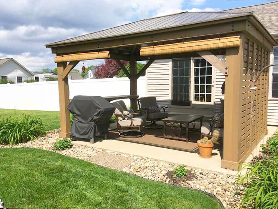 An outdoor living space is pictured on a partially cloudy day, featuring a wooden patio gazebo structure attached to the side of a house.

The gazebo has a dark brown slatted metal roof and sturdy brown wooden posts and beams. The sides facing the viewer are partially enclosed by vertical wooden slatted walls, and a bamboo roll-up blind is visible under the roof eave on the left side, offering adjustable shade.

The floor beneath the gazebo is a concrete patio, which hosts a comfortable outdoor lounge area. Furnishings include a covered barbecue grill on the far left, a dark brown outdoor rug anchoring the seating arrangement, and several cushioned swivel chairs and a low coffee table. A terracotta pot with a cactus plant sits near the right front post. Behind the seating area is a large glass window or door belonging to the adjacent house, which features light-colored horizontal siding.

The outdoor space is well-landscaped. In the foreground, there is a neatly manicured green lawn separated from the patio by a border of small river rocks and edging. Lush green plants and shrubs frame the immediate foreground and the left side of the structure.

Beyond the gazebo and to the left, a tall white vinyl fence separates the yard from a neighboring property, where a white house is visible under a blue and cloudy sky. The image successfully conveys a comfortable, well-equipped backyard space ideal for grilling and relaxation.