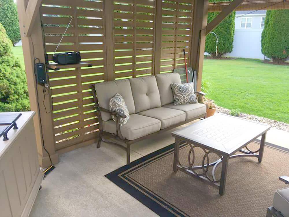 A medium-close indoor/outdoor shot captures a cozy seating area set up beneath a wooden gazebo or patio cover.

The primary focus is a three-seater outdoor sofa with light beige, thick cushions and throw pillows featuring a dark, patterned design. The sofa sits against a slatted wooden screen or wall, which provides privacy and shade. To the left of the sofa, a small black radio and a piece of electronic equipment are mounted to the slatted wall. A couple of garden tools are propped up near the right side of the sofa, next to a terracotta pot.

In front of the sofa is a rectangular coffee table with a light-colored, possibly glass or metal, top and an intricately designed metal base featuring interlocking ring patterns. The seating and coffee table are placed on an outdoor rug with a black and brown geometric border, defining the lounge area. The base of the structure is a smooth, light gray concrete patio.

Beyond the covered area, the image shows a neatly maintained green lawn framed by several dark green, conical shrubs or trees. A portion of the exterior of a light-colored house or structure is visible in the upper right background.

To the far left, a glimpse of a white kitchen counter or bar unit suggests an outdoor cooking or entertainment area is nearby. The overall atmosphere is one of relaxed, comfortable outdoor living, perfect for casual gatherings or listening to music.