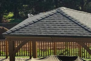 Timber gazebo with pyramid-style shingled roof on deck surrounded by trees, fence, and spotted dog peeking over top