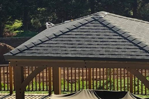 A photograph captures the upper portion of a wooden gazebo or pavilion structure, showcasing its roof and railing against a backdrop of trees on a sunny day.

The roof is prominently featured, characterized by a classic, multi-faceted pitched design, likely a hip roof. It is covered in dark gray or black asphalt shingles, laid in neat, overlapping rows. The ridge cap, running along the highest peak, is visible. Interestingly, a small, dark object with white spots, possibly a spotted stone or decorative finial, is resting on or built into the roof near the ridge on the left side. The roof overhangs the main structure slightly, finished with dark trim.

The main structure beneath the roof is built from light brown or medium-stained wood. Visible details include a thick horizontal header beam and an angled support brace beneath it, indicating a sturdy frame. This area appears to define a comfortable outdoor living space.

The enclosure beneath the header beam is finished with a wooden railing system. The railing consists of thin, vertical wooden spindles or balusters set closely together, providing safety and a degree of visual separation from the surrounding yard.

The gazebo is built on a deck or patio surface, visible below the railing. Part of the deck area is covered by a dark, possibly black, protective furniture cover in the foreground. Shadows cast by the railing indicate strong overhead sunlight.

In the background, the scene transitions immediately into a natural, wooded area. A dense mass of deep green trees and foliage fills the upper background. To the left, a mound of dark, loose material, possibly mulch, soil, or wood chips, suggests a landscaping project is ongoing nearby. The image conveys a sense of a finished, high-quality outdoor structure providing shelter and defining a specific activity area within a wooded backyard setting.
