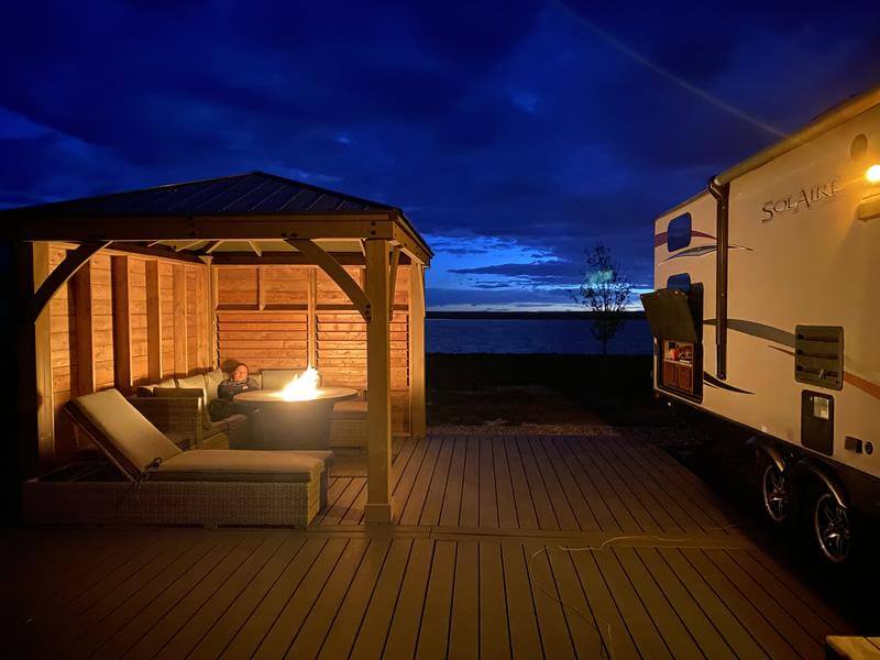 Twilight lakeside gazebo with fire pit, cozy seating, and SolAire RV parked nearby.