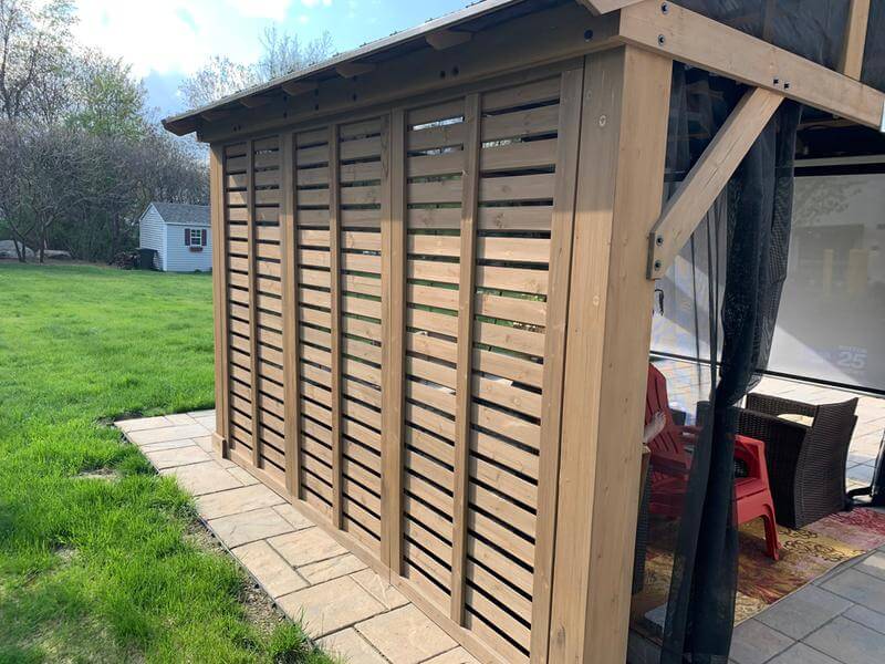 A close-up photograph captures the exterior of a light-toned wooden gazebo or covered patio structure on a bright, sunny day. The focus is on a solid wall of the gazebo, which is constructed of multiple vertical wood panels featuring horizontal slats or louvers. These slatted panels provide privacy and sun protection while still allowing air and some light to pass through. The wood is a natural, light brown color.

The gazebo wall rests on a rectangular border of flat, earth-toned pavers, which separates the structure from the surrounding bright green lawn. To the right, the open side of the gazebo is visible, revealing a glimpse of the interior. A corner post and the angled support beam of the roof are present, along with the edge of a dark, sheer mesh screen or curtain hanging down, likely for insect protection or added shade.

Inside the gazebo, on the flooring, a patterned rug in warm colors (reds and yellows) is visible, defining the seating area. A piece of red patio furniture, possibly a chair, is partially seen, alongside darker wicker or rattan furniture.

In the background, the scene transitions from the hardscaping to a well-maintained, slightly uneven green lawn. Further back, situated in a clump of trees and bushes, is a small, shed-like outbuilding. This shed has white siding, a dark roof, and a visible small window and door trim painted in a dark blue or black color. The surrounding mature trees and bushes are lush and green, framing the background of the image. The sunlight is strong, suggesting midday, casting clear light on the wood grain and the texture of the pavers. The overall image emphasises the detail and structure of the privacy wall and the transition from the manicured outdoor structure to the natural, grassy backyard.