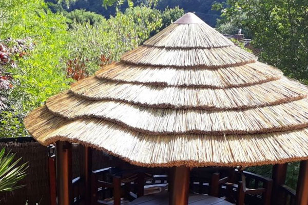 A bright, sunny, close-up photograph focuses on the distinctive, tiered roof of an outdoor tiki hut or thatched gazebo, suggesting a tropical or exotic setting.

The most prominent feature is the roof, which is made of thick, natural straw or reed thatch. The thatch is applied in multiple, layered tiers, creating a dramatic, stepped cone shape that rises to a central peak. The layers cast deep shadows on one another, emphasizing the texture and depth of the material. The straw is a warm, golden-brown color, catching the sunlight beautifully. At the very peak of the roof, a small, dark conical cap finishes the structure. The substantial overhang of the thatch provides significant shade.

Below the thatched roof, the supporting structure of the gazebo is partially visible. The vertical posts and beams are made of dark-stained or naturally dark wood, contrasting sharply with the lightness of the thatch. The interior space glimpsed beneath the eaves shows dark wood railing or seating and a light-colored surface, possibly a round table. To the left, the dark frame supports a section of fencing or siding made of woven bamboo or reeds. A piece of a lush, broad-leaved palm-like plant is also visible near the frame.

The background is dominated by a dense and vibrant green landscape. Numerous trees and bushes with rich, varied green foliage fill the scene, confirming the tropical, garden, or heavily planted outdoor environment. The bright, high-contrast sunlight suggests midday, illuminating the golden color of the thatch and the deep shadows underneath the canopy. The overall image evokes a strong feeling of relaxation, shade, and a vacation-like atmosphere, ideal for an outdoor dining or lounge area.