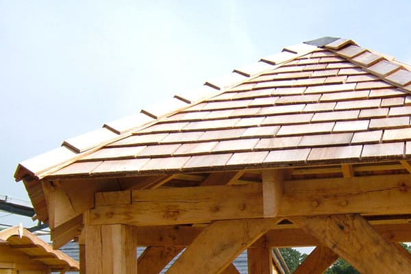 A close-up architectural photograph focuses on the roof and structural joinery of a timber-framed gazebo or outdoor pavilion, emphasizing traditional craftsmanship. The image is taken on a bright day against a pale sky.

The roof is the most prominent feature, covered in overlapping wooden shingles or shakes that display a warm, natural light brown to golden color. The clean, defined edges of the individual shingles are clearly visible, suggesting they are new or well-maintained cedar or similar wood. The roof is steeply pitched, with the front edge (eaves) overhanging the main structure.

The supporting structure beneath the roof is constructed from heavy, rough-hewn timber, characteristic of traditional framing techniques like post-and-beam. The wood is a deep golden-brown, possibly oak or an older, cured softwood, and exhibits natural grain, knots, and slight texture, unlike the smooth modern wood in other images. Visible details include a thick horizontal tie beam, an upright support piece directly beneath it, and an angled support beam—or brace—extending downwards to the right (the corner post itself is mostly obscured). The timber connections appear robust, using classic joinery techniques, though specific fasteners are not clearly visible.

The underside of the roof eaves is visible, showing the wooden decking and rafters that support the shingles. The framing elements beneath the eaves are substantial and functional.

In the lower left corner, a portion of a smaller, similar-style structure is partially visible, suggesting this is part of a larger complex or outdoor display. The background is mostly a pale, overcast or bright white-blue sky, which provides a high-contrast backdrop that accentuates the warm color and texture of the wood shingles and beams. The overall impression is one of timeless, durable, and rustic outdoor construction, prioritising natural materials and substantial timber framing.