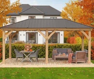 Wooden gazebo with a black metal roof and string lights, featuring a dining table with pumpkins and a lounge area with cushioned seating, set on a wooden deck in a landscaped garden with autumn trees and a white house in the background.