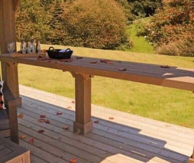 Outdoor wooden deck with a long table holding glass bottles, a wine glass, and a black basket, surrounded by scattered autumn leaves and a grassy garden backdrop.