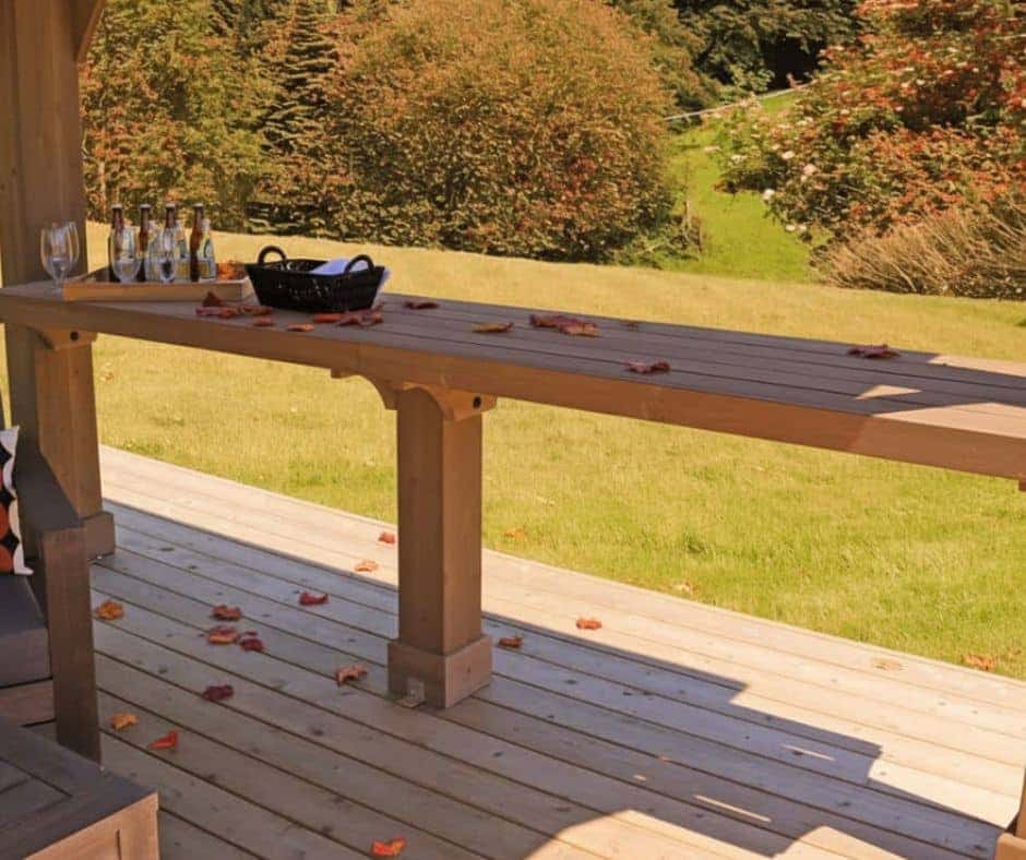 Outdoor wooden deck with a long table holding glass bottles, a wine glass, and a black basket, surrounded by scattered autumn leaves and a grassy garden backdrop.
