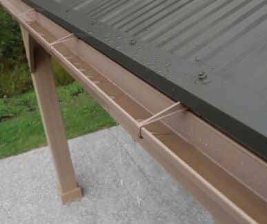 Close-up of a dark corrugated metal roof with water droplets and a brown gutter system dripping rainwater, set against green grass and concrete ground.