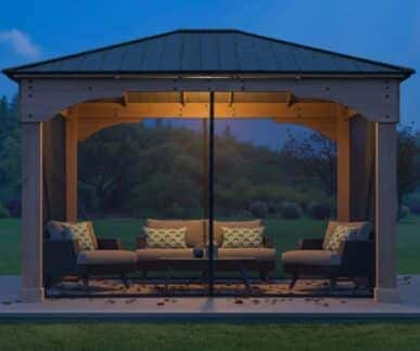 Outdoor gazebo with a dark metal roof and wooden frame, illuminated by a central light, featuring cushioned seating and a coffee table, with autumn leaves scattered on the floor.
