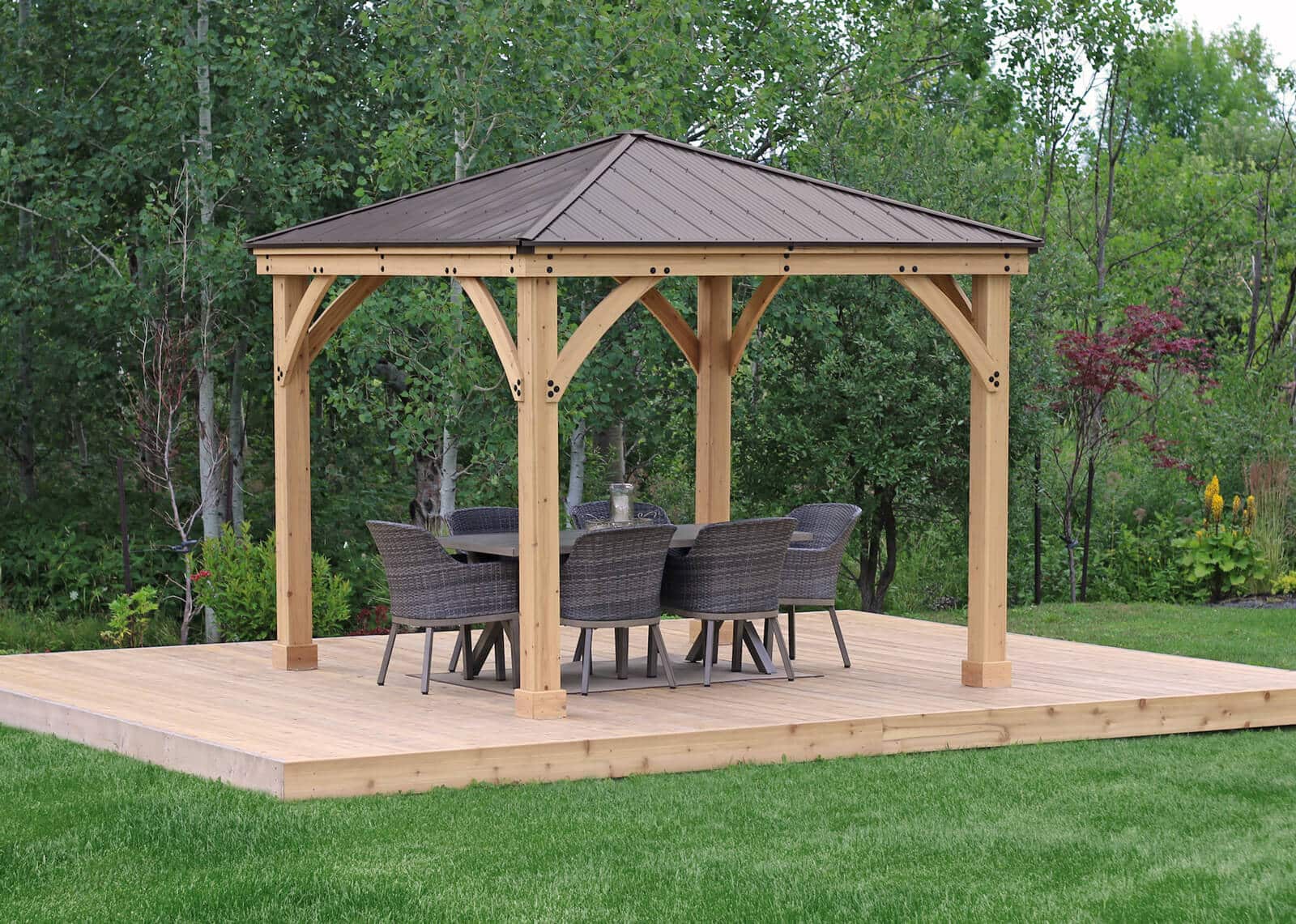 Wooden gazebo with metal roof on a raised platform, featuring a dining table and six wicker-style chairs in a grassy garden surrounded by trees.