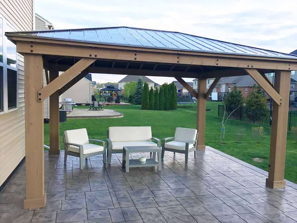 A Yardistry cedar wood gazebo with aluminium roof built on to a grey paved area, with comfortable sofa and chair seating arranged around a small table.  The garden is lush with green grass and trees in the background, plus a patio and bbq area for outdoor living.