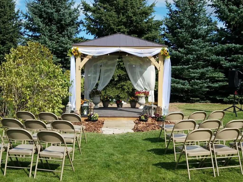 An outdoor wedding ceremony setup. In the center, there is a wooden gazebo or arbor with a dark, peaked roof, draped with sheer white fabric curtains. The gazebo is decorated with small bunches of sunflowers or yellow flowers at the top corners and contains a raised wooden platform and some decorative planters and lanterns.

In front of the gazebo, arranged on a green lawn, are two curved rows of beige/tan folding chairs, set up for guests. A small stone pathway lined with wood chips leads from the seating area up to the gazebo. The backdrop consists of a dense line of tall evergreen trees and bright green bushes under a sunny sky.