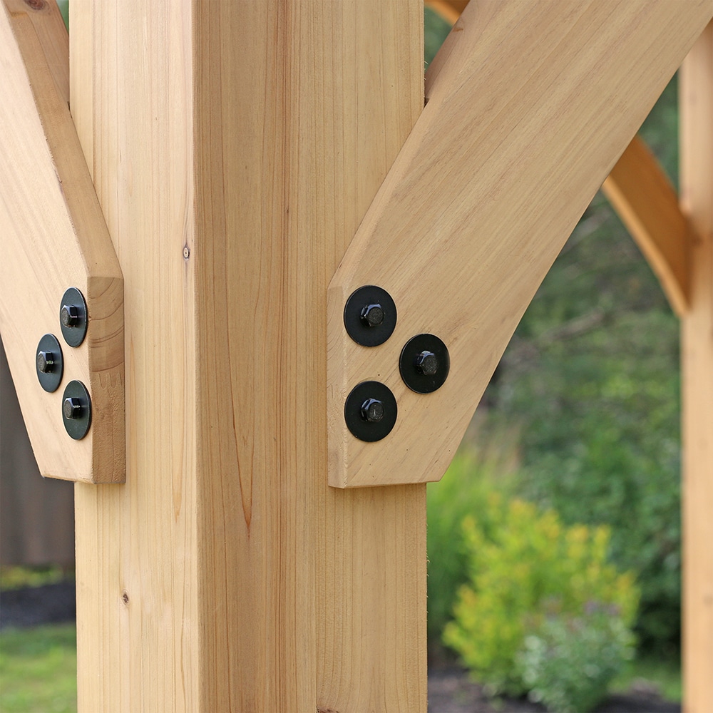 The image displays a close-up of a wooden structure, likely a gazebo, pergola, or post-and-beam construction, focusing on a joint.

Here is a detailed alt text description:

Close-up of a timber frame joint showing two diagonal wooden braces fastened to a vertical post. The wood is light-colored and freshly milled. Each brace is secured with three matte black circular fasteners, which appear to be large washers covering bolt heads or lag screws, contrasting sharply against the light wood. The structure is set outdoors with blurred greenery visible in the background.