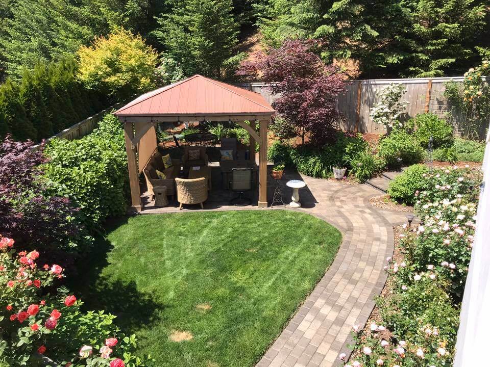 An elevated view of a meticulously landscaped backyard garden and patio area. In the center, a light brown wooden gazebo with a reddish-brown metal hip roof sits on a paved patio section. Outdoor furniture, including wicker chairs, is visible inside the gazebo.

The yard features a small, neatly maintained patch of green lawn bordered by a curving path made of interlocking light brown pavers. The path is surrounded by various plantings, including bushes with dark red and yellow leaves, and climbing roses with pink and red blooms in the foreground. A white bird bath stands next to the gazebo. The perimeter of the yard is enclosed by a wooden fence, and the background rises sharply into a densely forested hill of evergreen trees.