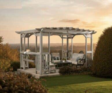 Wooden pergola with a slatted roof, featuring cushioned armchairs and a loveseat arranged around a coffee table, with a side table holding a cake and teapot, set in a landscaped garden with trimmed bushes and a scenic sunset view over distant fields and trees.
