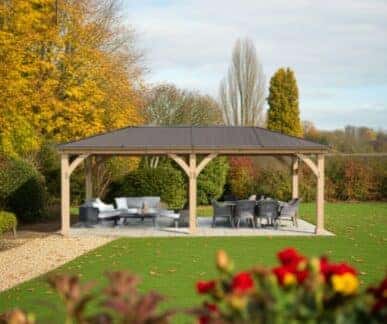 Large wooden gazebo with a dark metal roof, featuring cushioned chairs and sofas arranged around tables on a paved patio, surrounded by a green lawn, red flowers, and trees with vibrant autumn foliage.