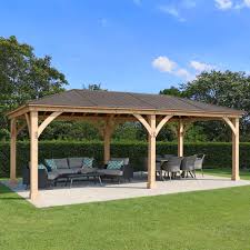 Large wooden gazebo with dark shingle roof on a concrete pad, featuring a sectional sofa, coffee table, and dining set, surrounded by grass and trees in a landscaped garden.