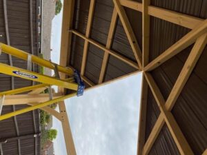Partially constructed wooden roof with metal sheets and triangular truss beams, yellow Werner ladder positioned underneath in a fenced residential garden.