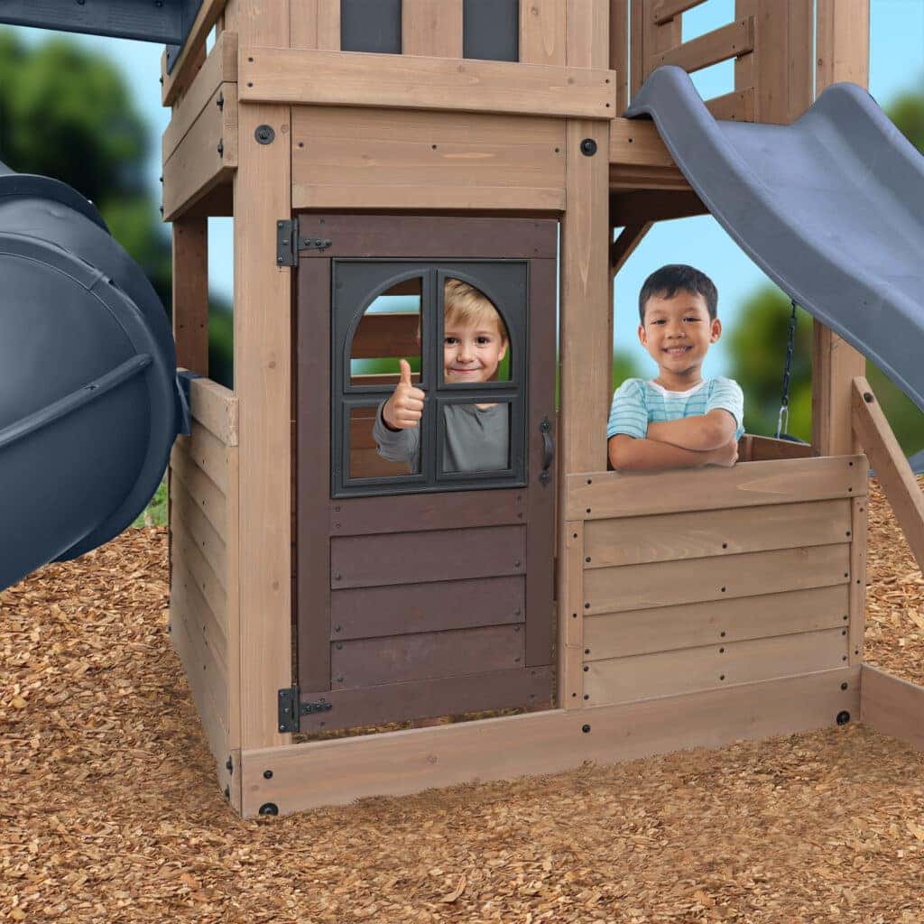 This image features two boys playing in the ground-level section of a wooden playset.

Here is a detailed description:

Playset Structure: The structure is made of light brown, stained wooden planks forming an enclosed area that resembles a small playhouse or fort.

Door: The front of the playhouse has a dark brown door with a decorative window pane (possibly plastic).

Window/Opening: To the right of the door, there is an open window or cutout above a wooden panel barrier.

Slides: Parts of two different slides are visible: a grey tube slide on the far left and the top of a standard grey straight slide on the far right.

Children:

Boy 1 (in doorway): A boy with light hair is framed in the window of the dark brown door. He is giving a thumbs-up sign with one hand and smiling.

Boy 2 (in window): A second boy is standing in the open window area, smiling with his arms crossed over his chest.

Ground Cover: The playset is installed over a bed of wood chips or mulch.

Background: The background is blurred, showing green foliage and a clear blue sky, indicating a bright, outdoor setting.

The image conveys a sense of friendship and fun in the backyard.